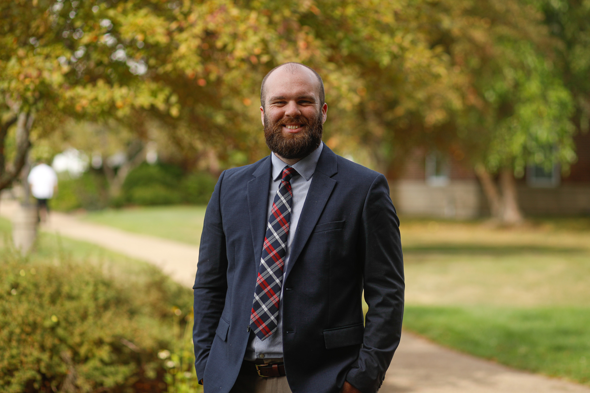 A man with a beard wearing a suit and plaid tie stands outside on a path, with trees and greenery in the background.