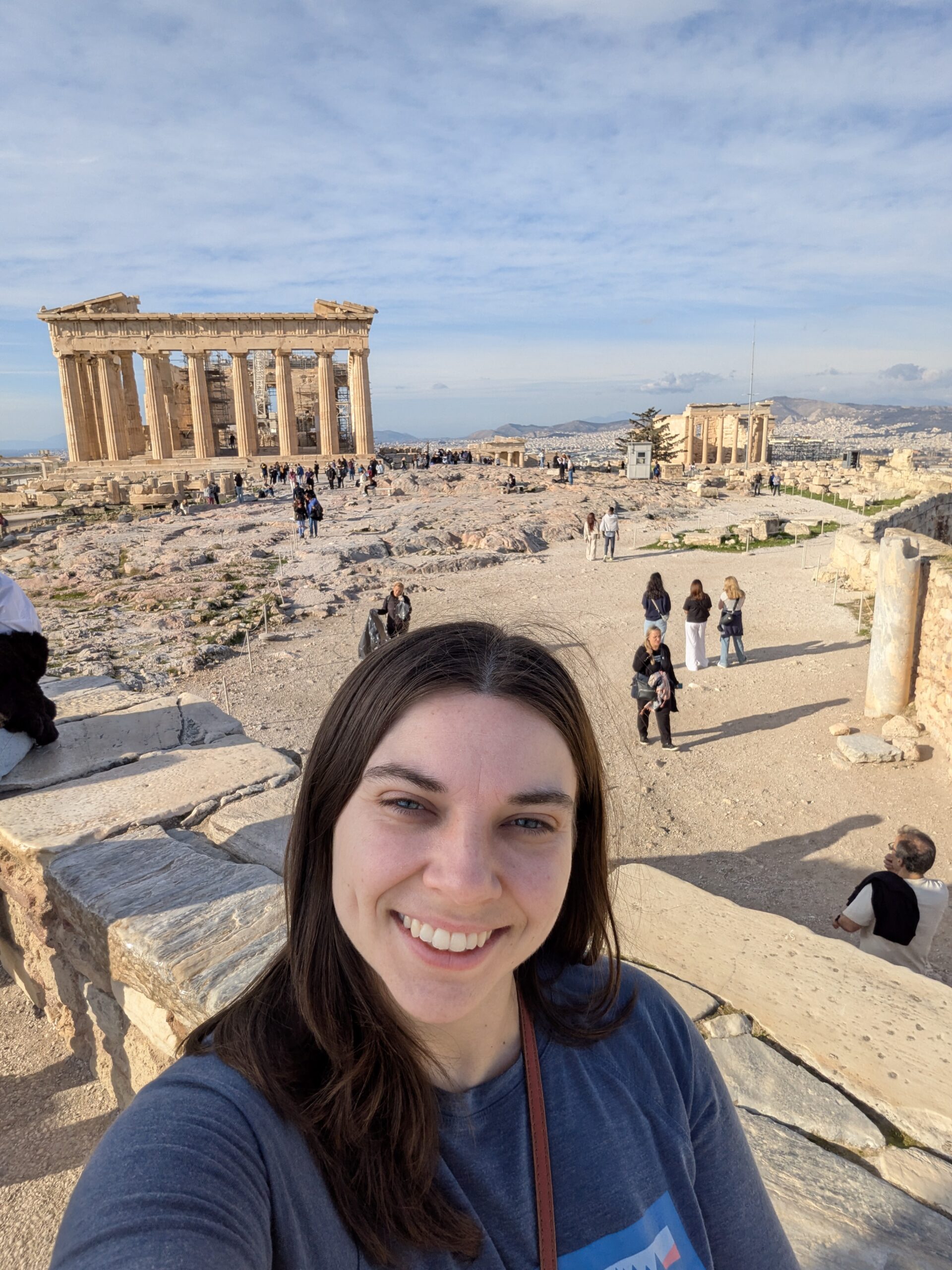 A woman takes a selfie at the Acropolis with the Parthenon in the background. Several other visitors are visible at the historic site under a partly cloudy sky.
