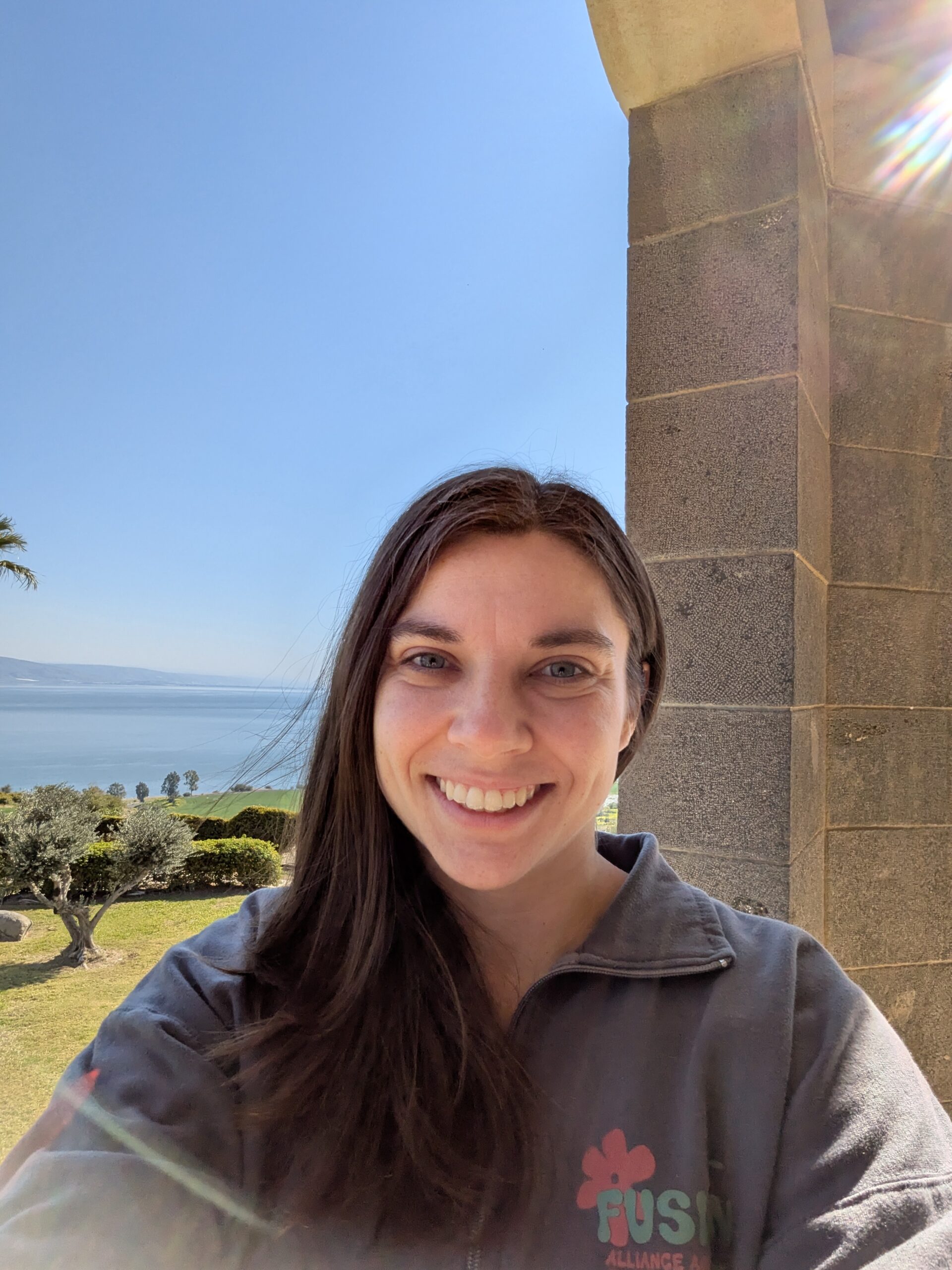 Woman with long brown hair smiling, taking a selfie outdoors near a stone pillar with a scenic background of trees, grass, and a large body of water under a clear blue sky.