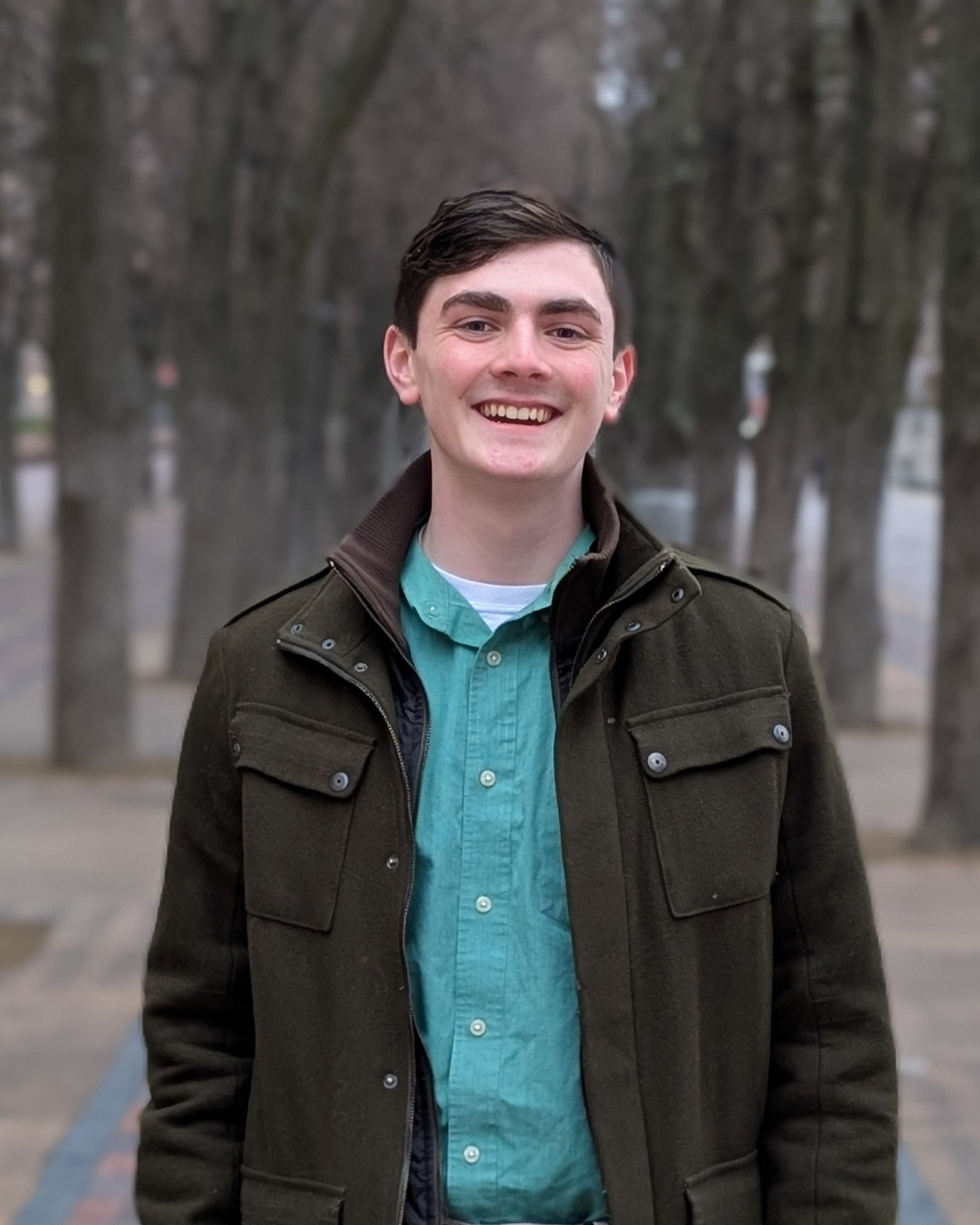 A young man with short brown hair is smiling and standing with his arms crossed in front of a rocky background. He is wearing a white sweater and a black collar shirt.