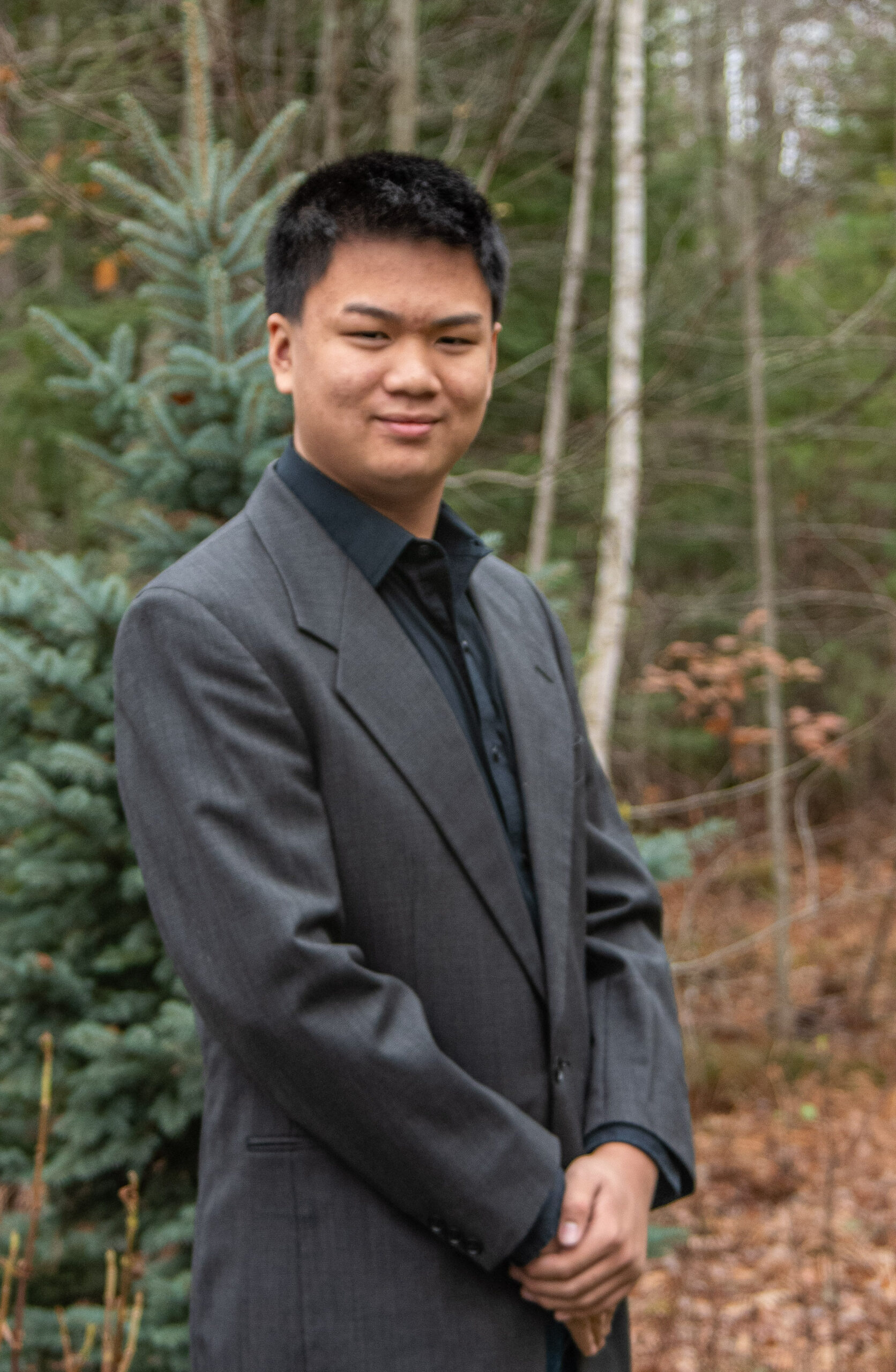 A young man with curly hair smiles while leaning on a wooden railing, with greenery and rocks in the background. He wears a light blue polo shirt.