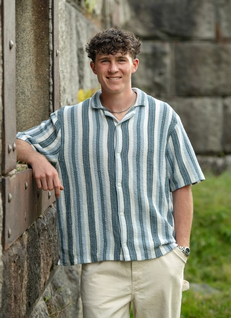 A young person with wavy hair wearing a light blue shirt and beige pants sits on rocks by the sea, with a calm expression and the ocean in the background.