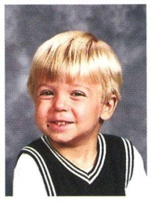 A black and white photo of a young child with shoulder-length hair and bangs, smiling at the camera, wearing a dark sweater with white diamond patterns.