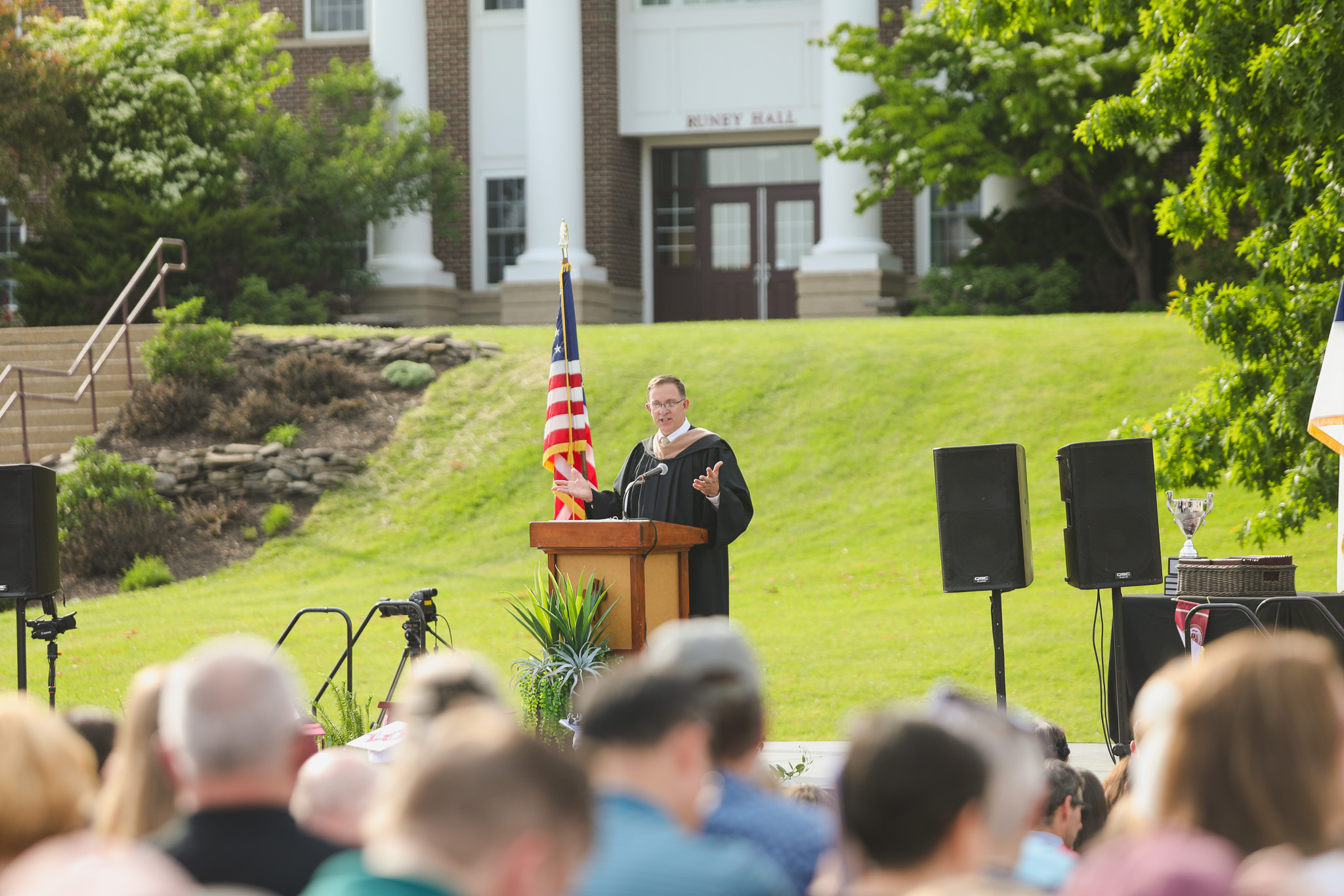 A graduate in a white cap and gown stands at a podium, speaking into a microphone on a stage decorated with plants.