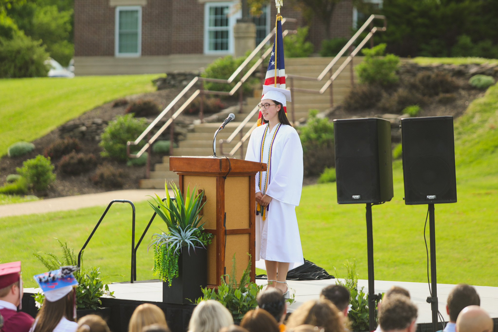 A graduate in a white cap and gown stands at a podium, speaking into a microphone on a stage decorated with plants.