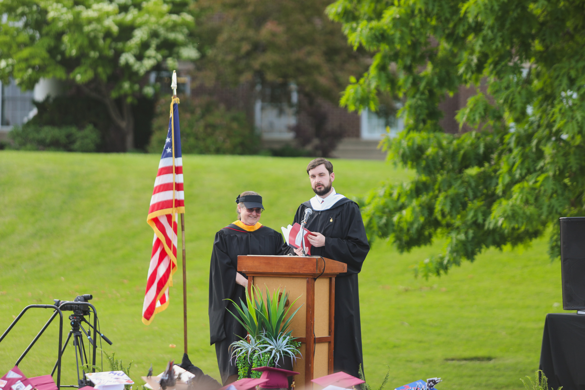 A graduate in a maroon cap and gown smiles while holding up a medal. He is wearing multiple honor cords and a medal around his neck.