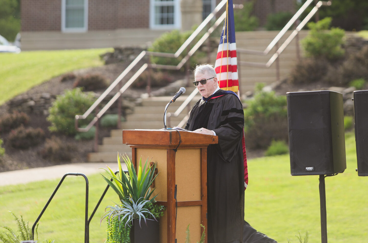 A person in a graduation gown stands at a podium on a stage, speaking into a microphone, with plants and black curtains in the background.