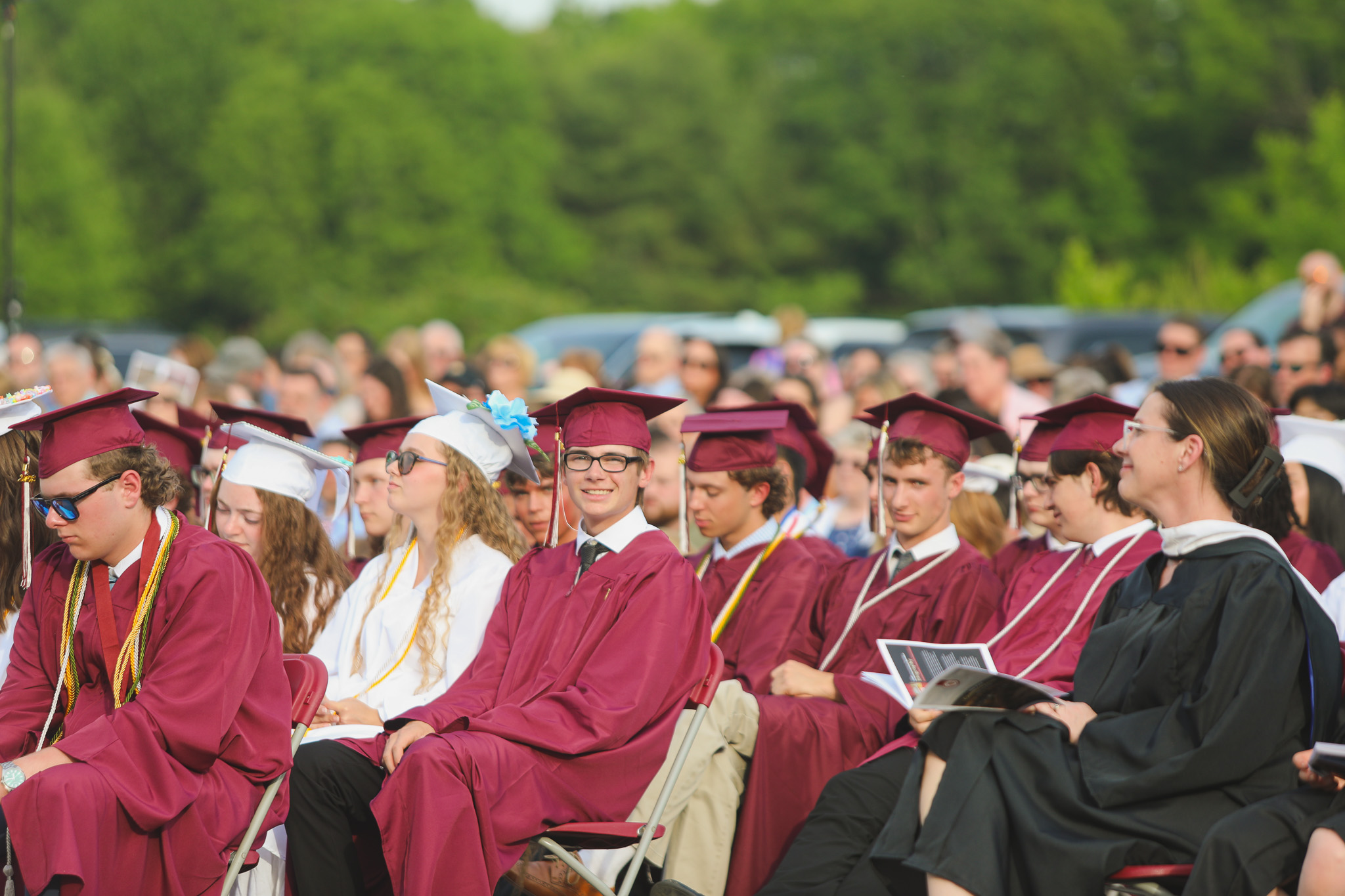A graduate in a maroon cap and gown is speaking at a podium outdoors, with a yard in the background and a white flag beside him.