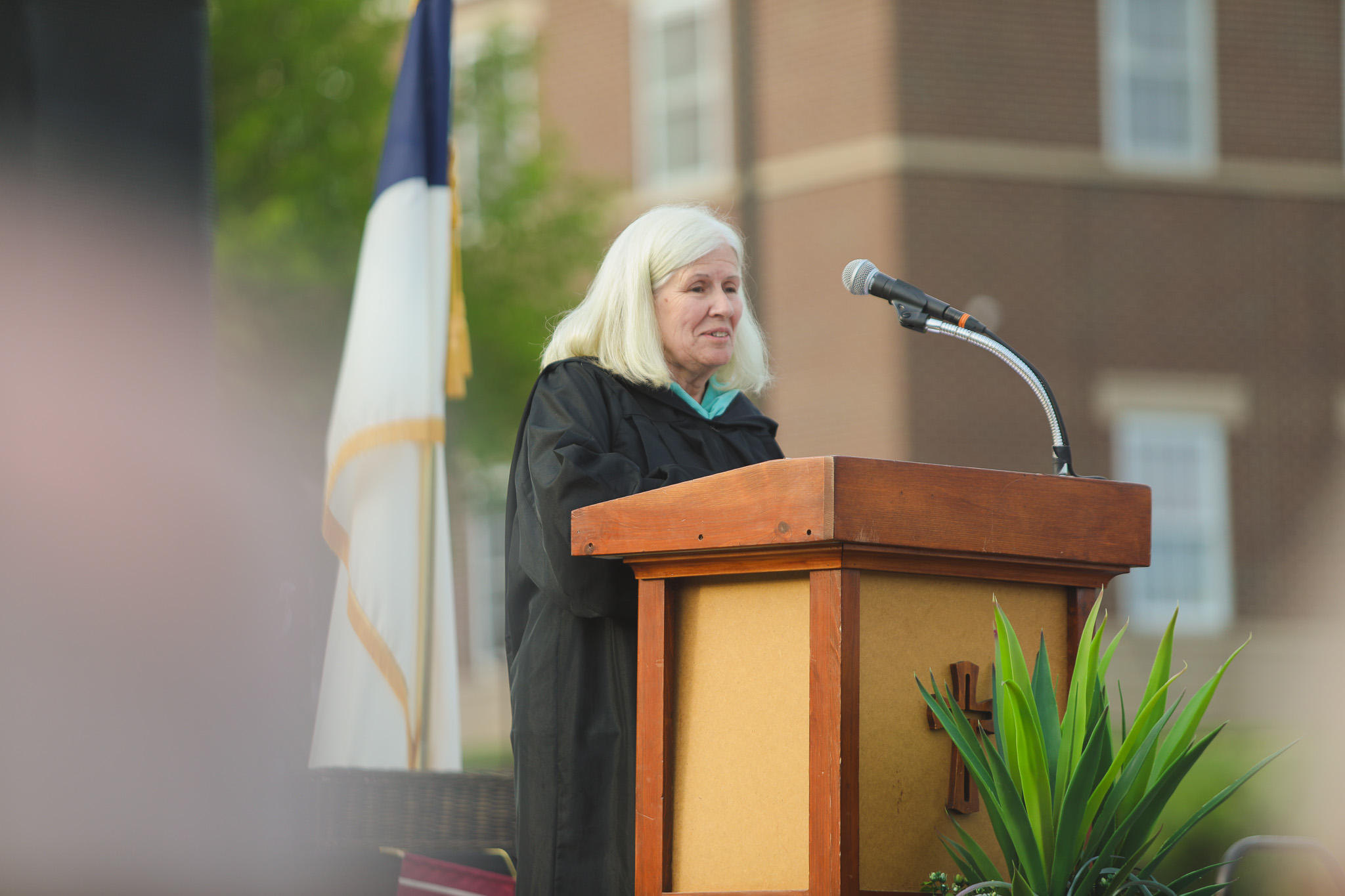A person in a graduation gown stands at a podium on a stage, speaking into a microphone, with plants and black curtains in the background.