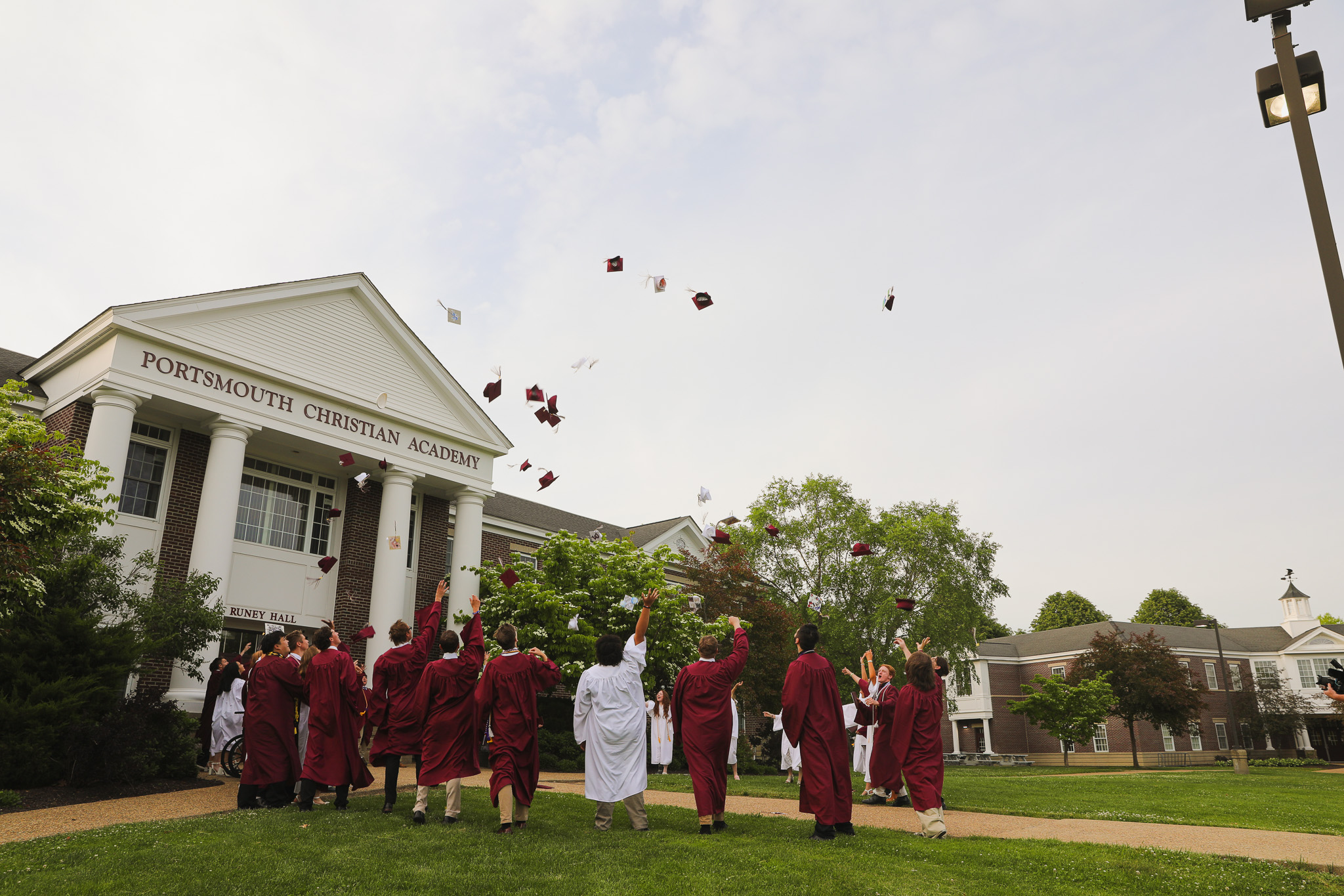A line of students in graduation robes and caps walk down steps outside, with green bushes and a building in the background.