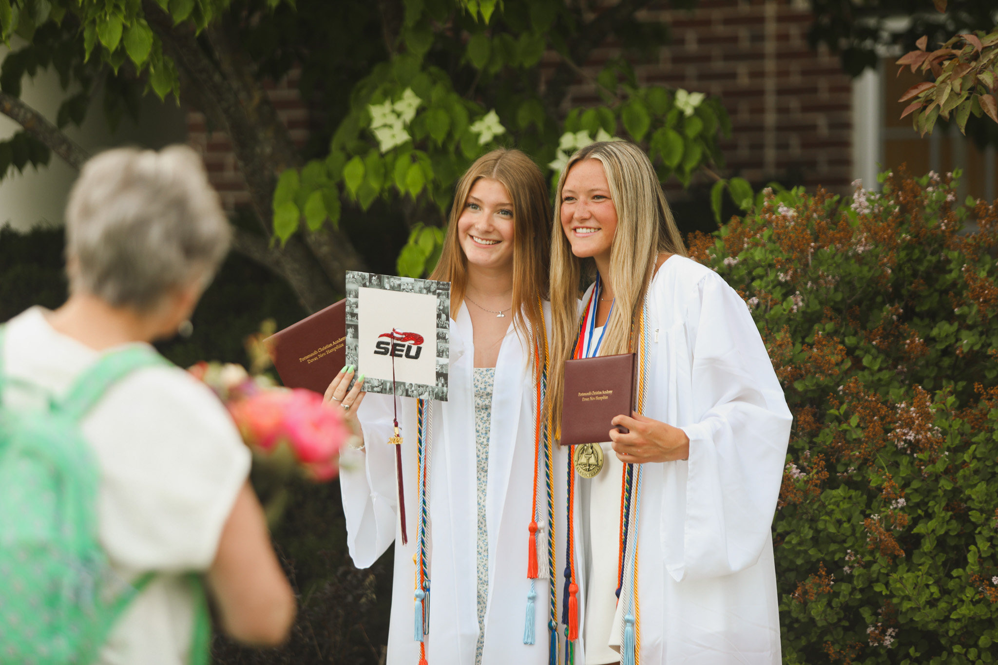 A person in a white graduation gown and cap stands outdoors on a grassy area, smiling. Another person in a similar outfit is seen in the background.