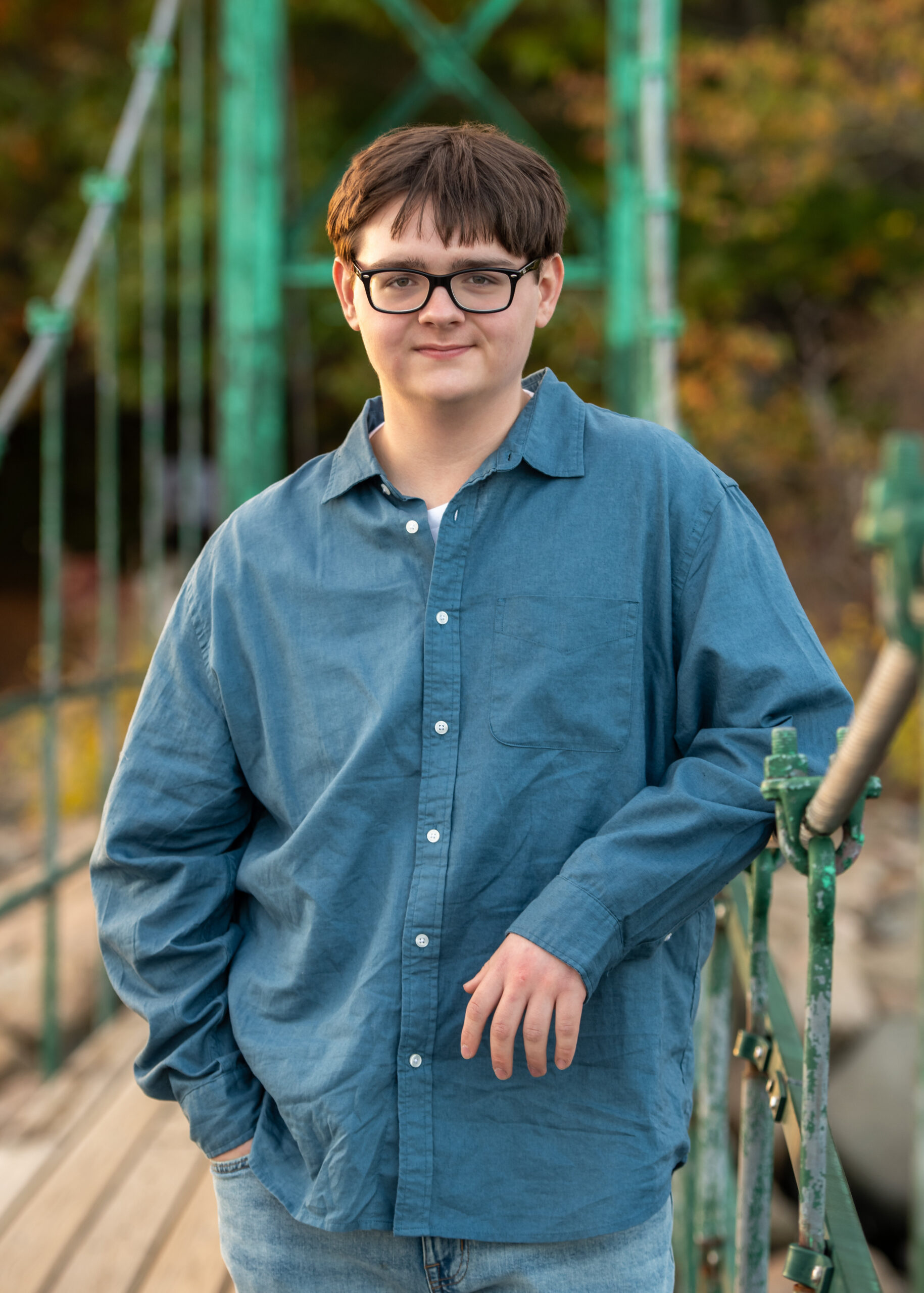 A young man in a blue polo shirt stands outdoors, leaning against a tree and smiling. The background is blurred with warm sunlight filtering through out-of-focus trees.